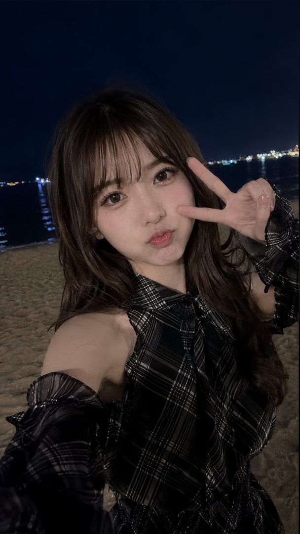 Young woman with long brown hair taking a playful nighttime selfie on the beach while making a peace sign.