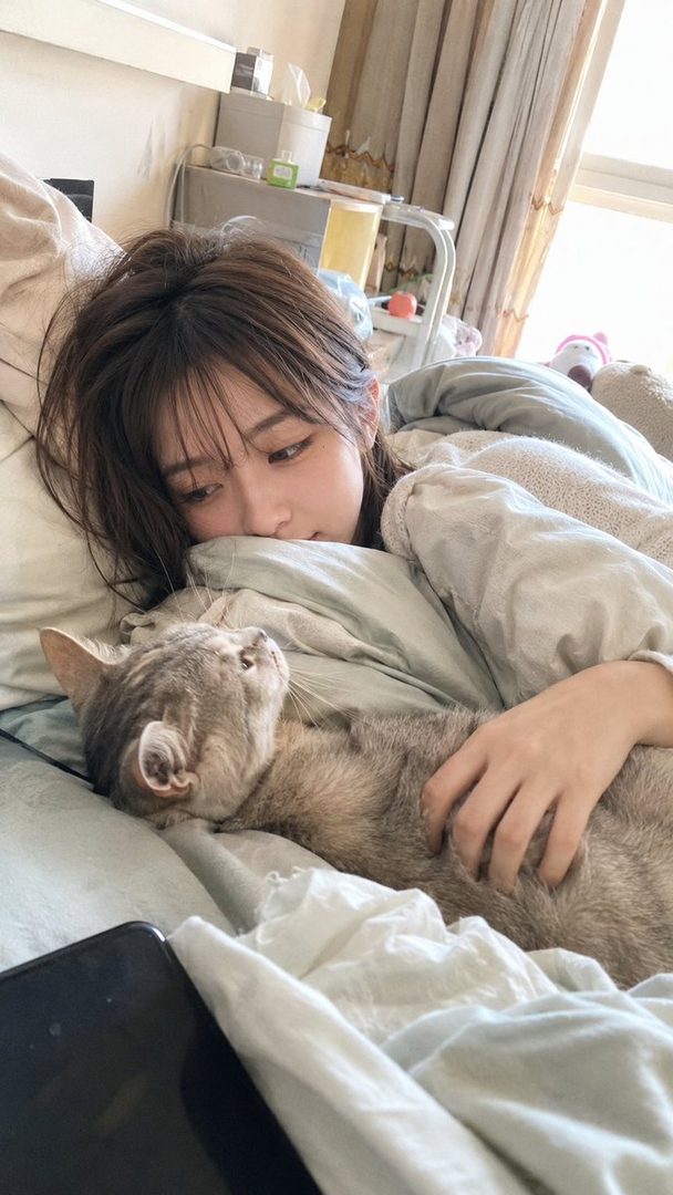A young woman resting in bed cuddling her gray cat on a lazy Sunday morning.