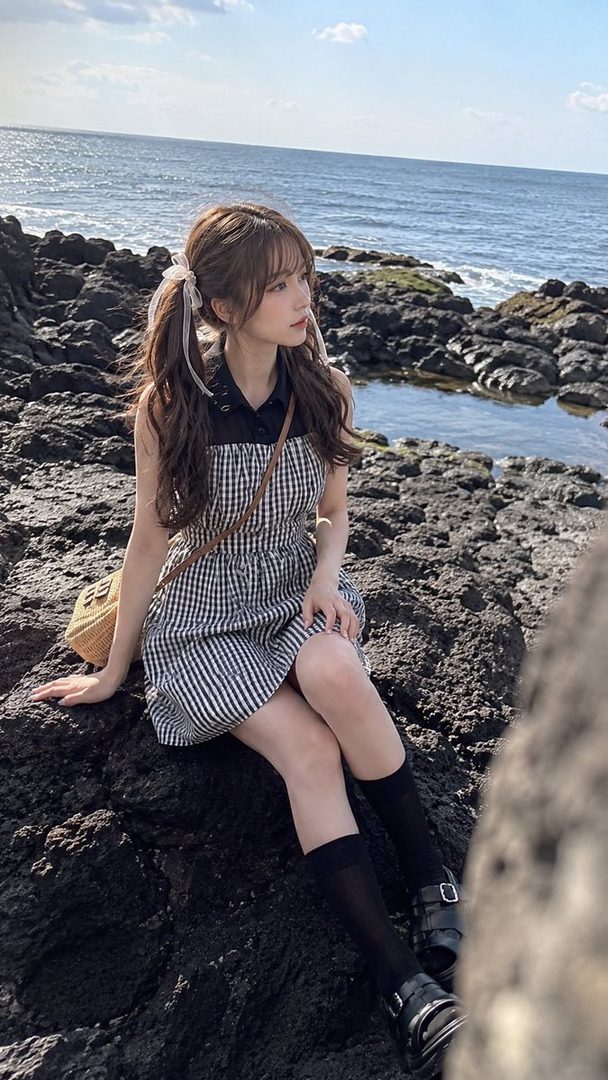 Young woman in a gingham dress sitting on rocky ocean shore under bright daylight.