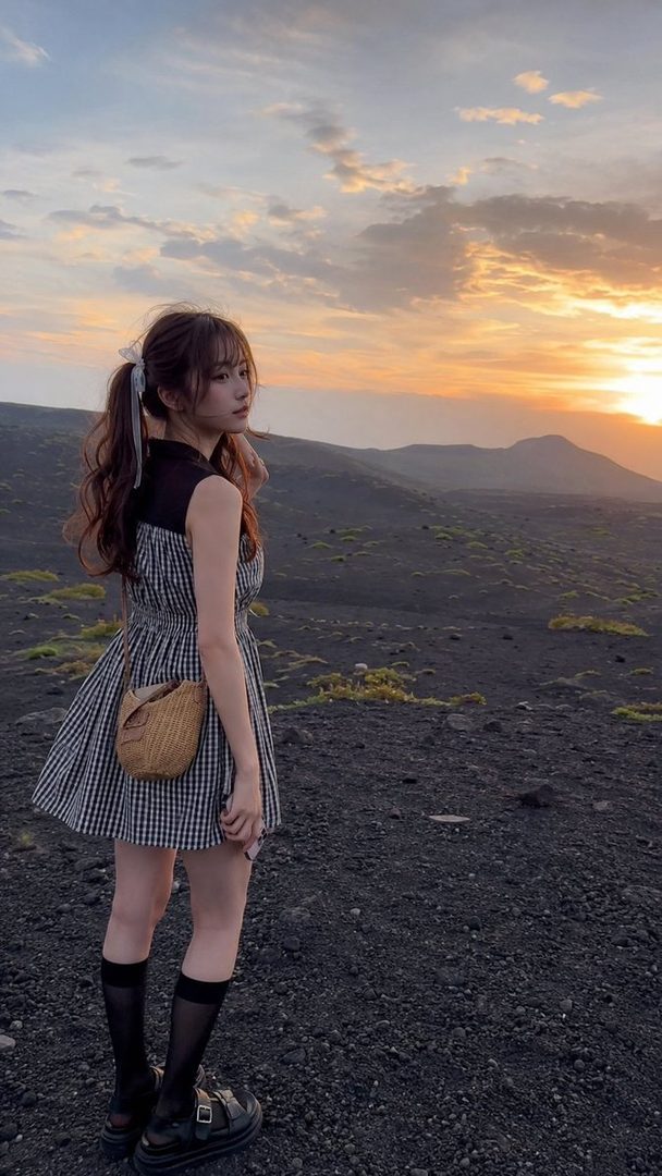 Young woman in a gingham dress with ponytail standing on volcanic rocks at sunrise.