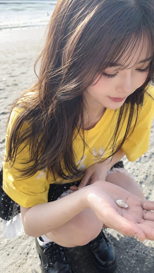 Young woman with long brown hair wearing a yellow t-shirt crouches on a sunny beach and examines a small seashell in her hand.