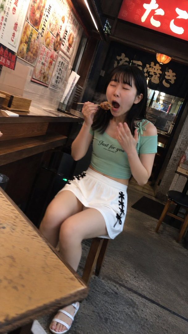 Young Japanese woman eating takoyaki at a food stall with eyes closed and playful expression.