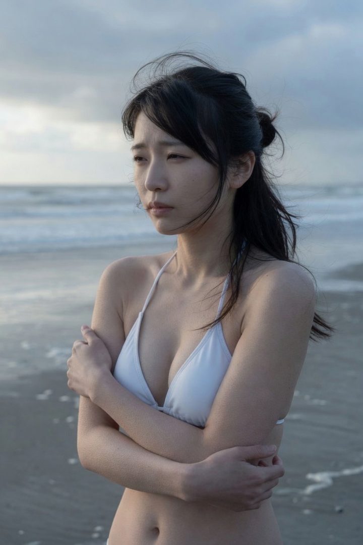 A young East Asian woman in a white bikini stands on a windswept beach at dusk with arms crossed, gazing pensively to the side.