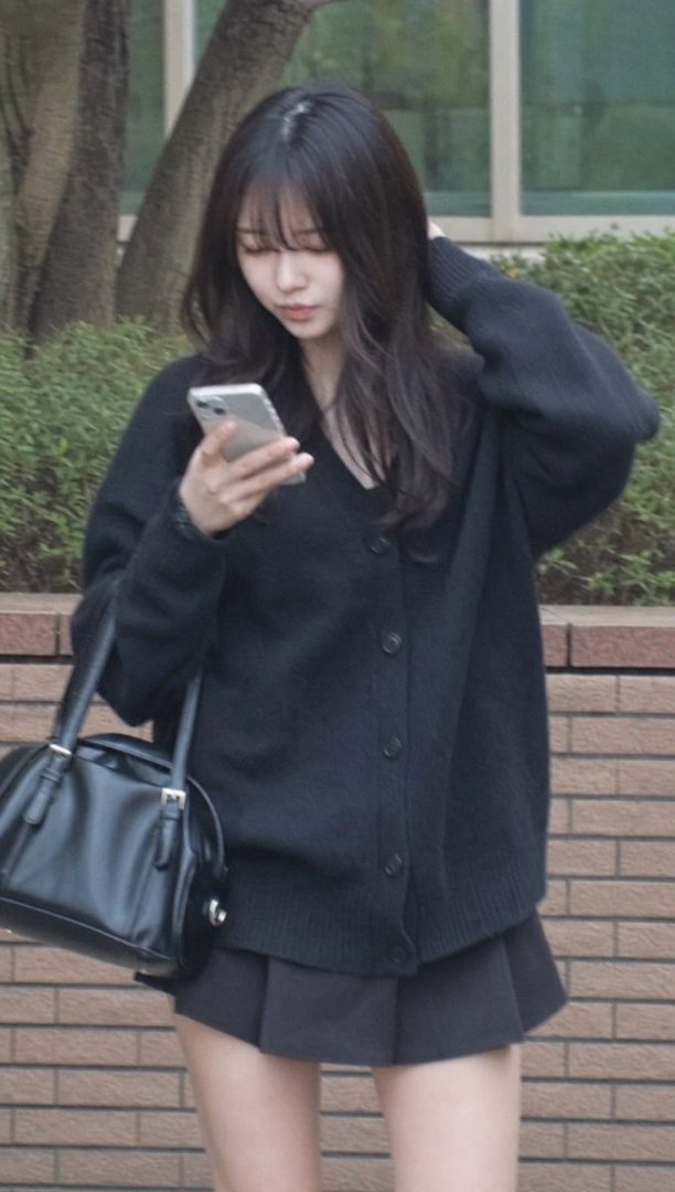 A young East Asian woman with long black hair checks her smartphone while standing outdoors in casual black knitwear and a pleated skirt.