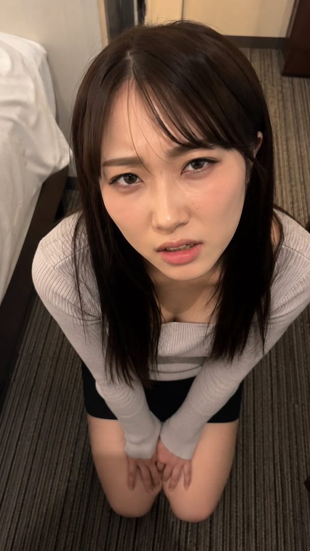 Young East Asian woman with long dark brown hair and bangs wearing a light gray sweater, kneeling on carpet in a bedroom and looking up at the camera.