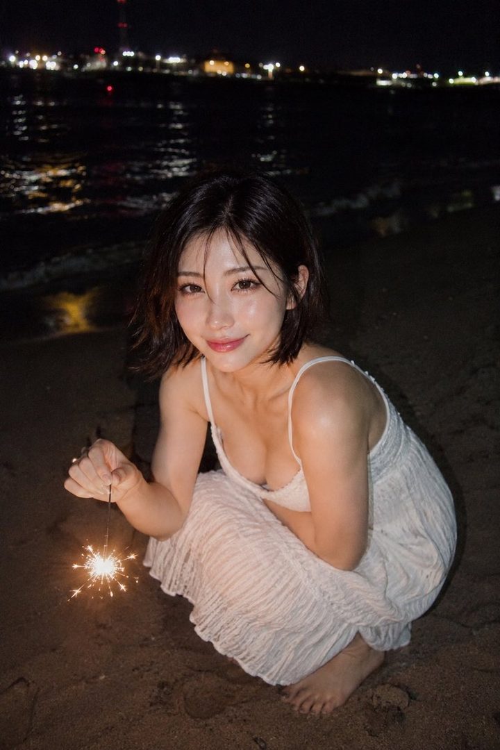 A young East Asian woman in a white dress crouches on a dark sandy beach at night, smiling while holding a lit sparkler with city lights across the water behind her.