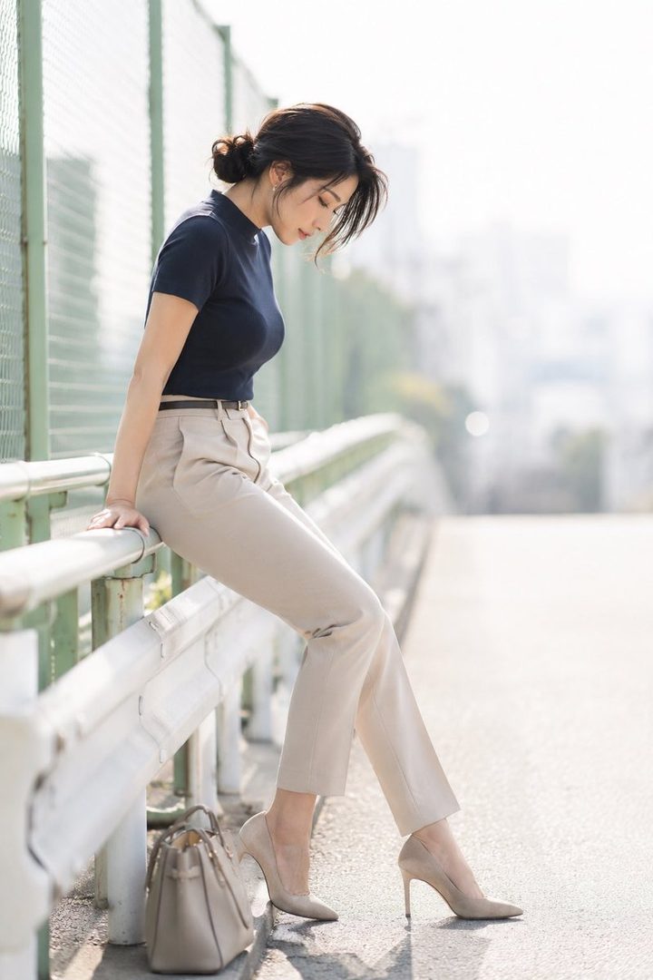 Elegant woman with dark hair in an updo leaning on a city railing, wearing a navy top, beige trousers and high heels with a handbag nearby.
