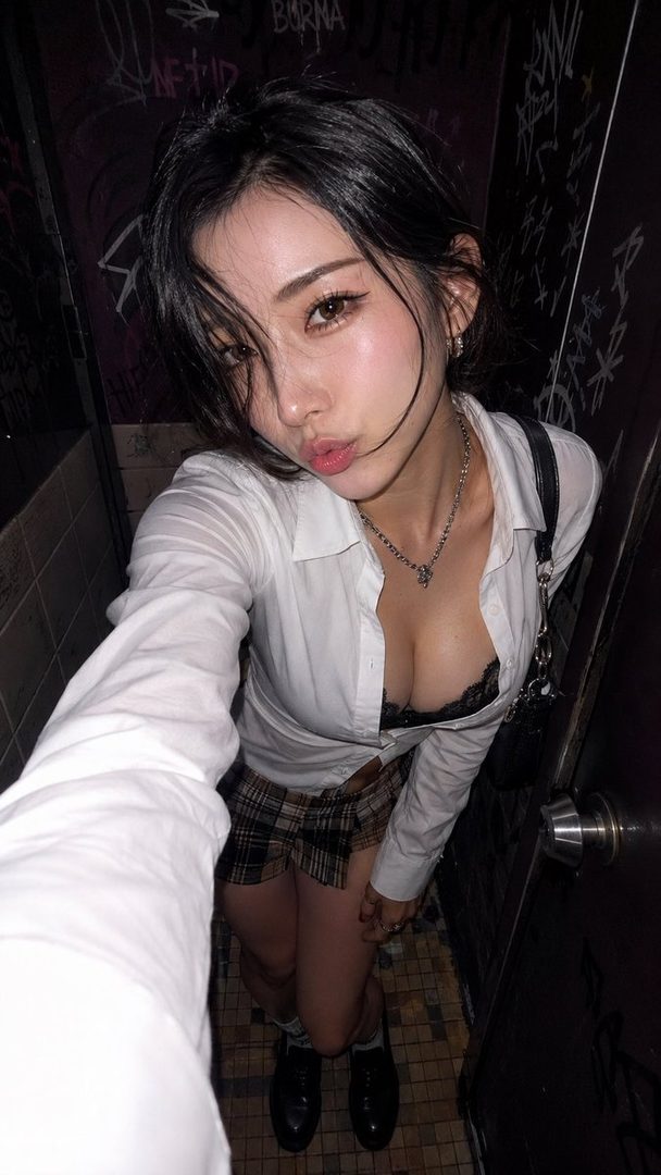 A young East Asian woman taking a selfie in a dimly lit graffiti-covered bathroom stall, wearing an unbuttoned white shirt revealing a black bra and a short plaid skirt.