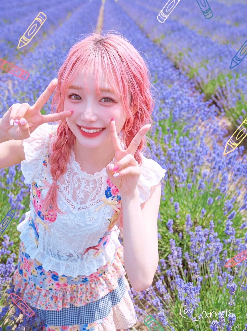 Young woman with pink braided hair smiling and flashing peace signs in a blooming lavender field under bright daylight.