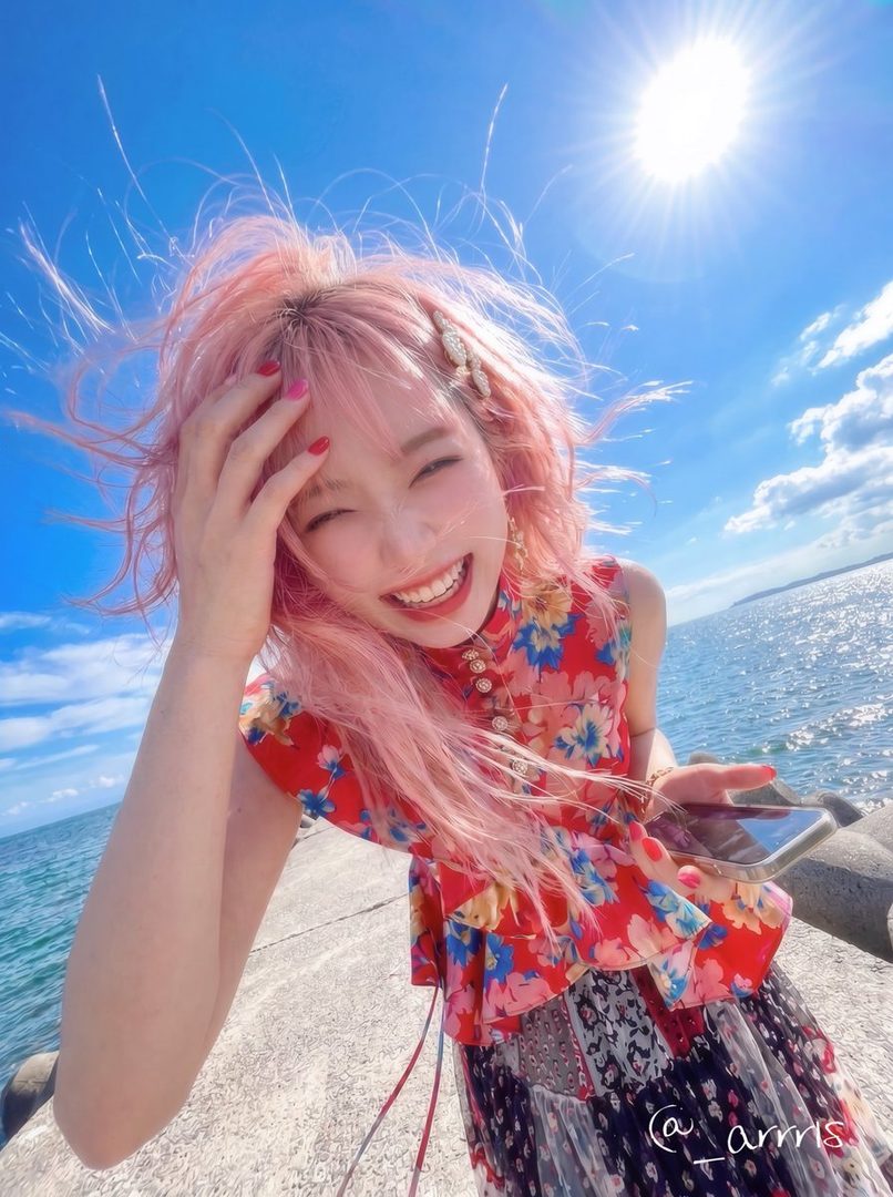 Joyful young woman with wind-swept pink hair taking a sunny selfie by the ocean on a bright summer day.