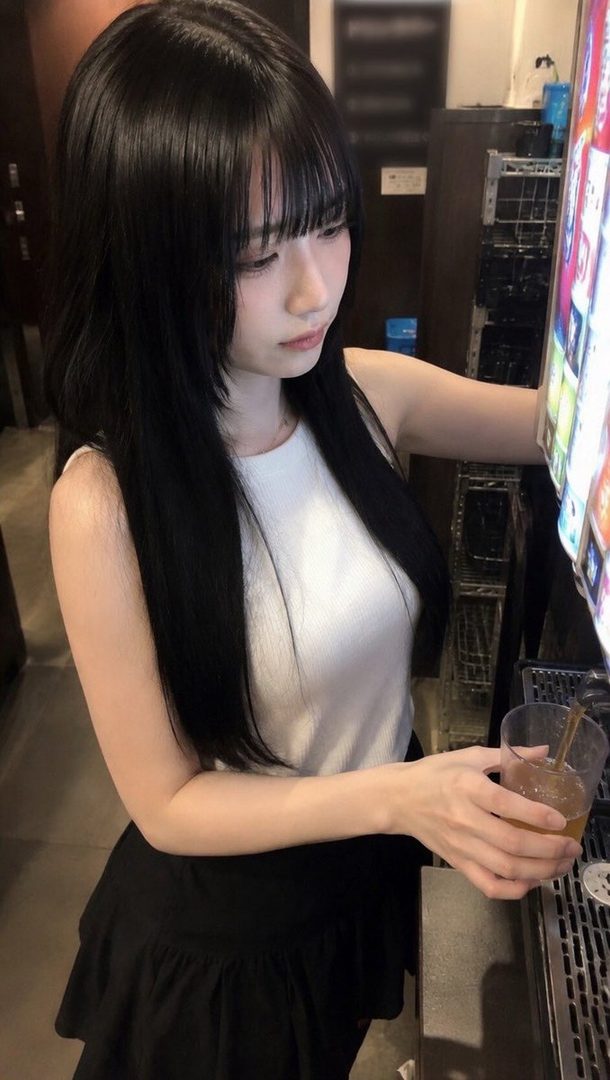 Young Japanese woman with long straight black hair and bangs, wearing a white sleeveless top and black skirt, standing indoors at night holding a drink.
