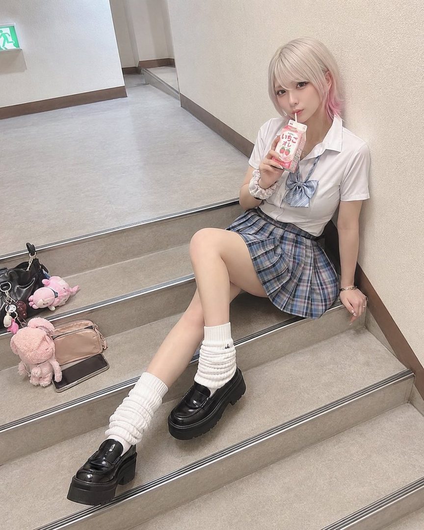 Young woman with pastel pink-tipped hair wearing a Japanese school uniform sits on indoor stairs sipping a juice box.