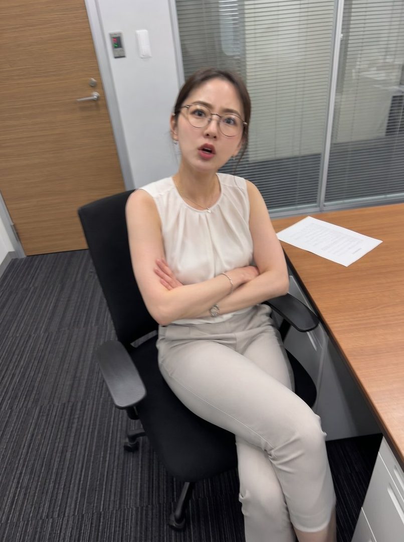 Professional woman with glasses sitting in an office chair with arms crossed, wearing a sleeveless blouse and trousers.