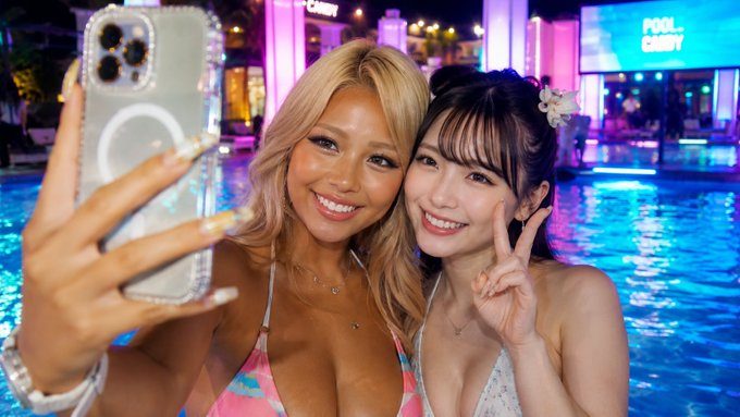 Two young Japanese women taking a joyful selfie together at a neon-lit night pool, one with blonde hair and the other with dark hair and a flower accessory.
