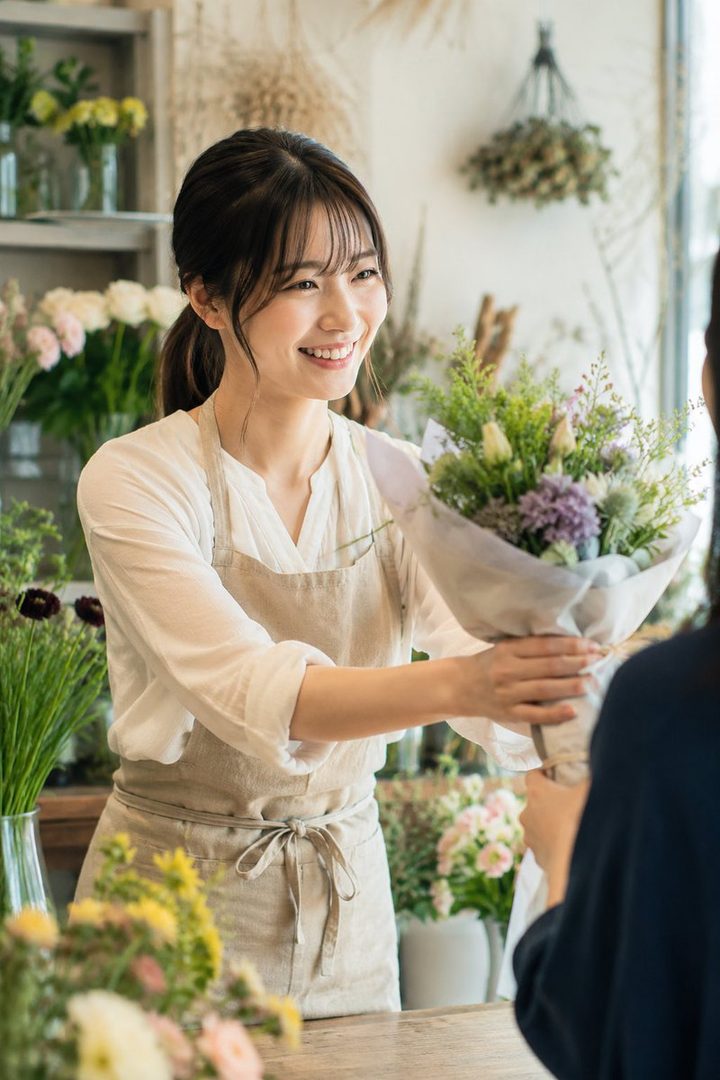 A smiling young Japanese woman in a floral apron handing a fresh bouquet to a customer inside a bright flower shop.