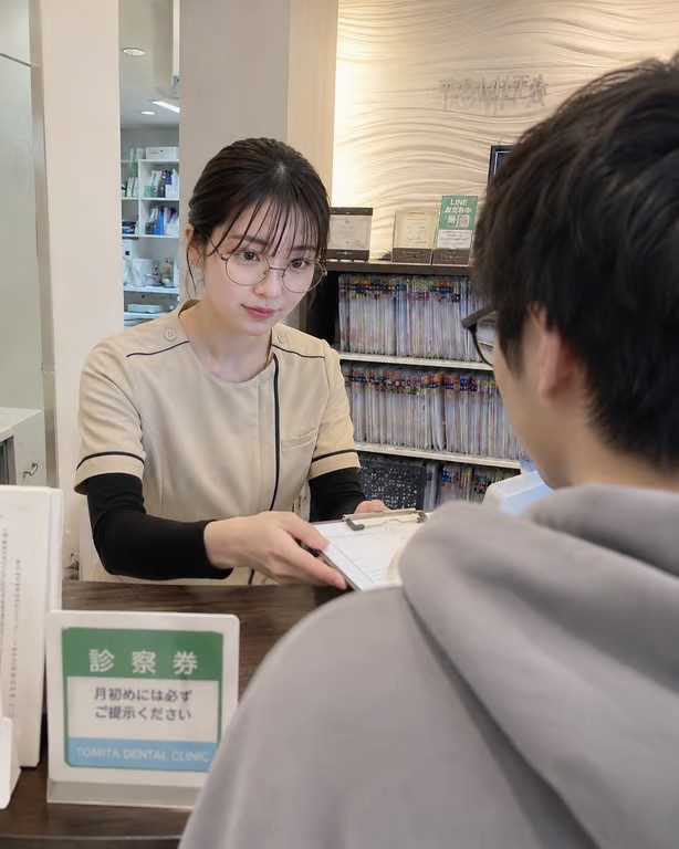 A young Japanese woman with glasses and dark hair in a dental clinic uniform hands a clipboard to a patient at the reception desk.