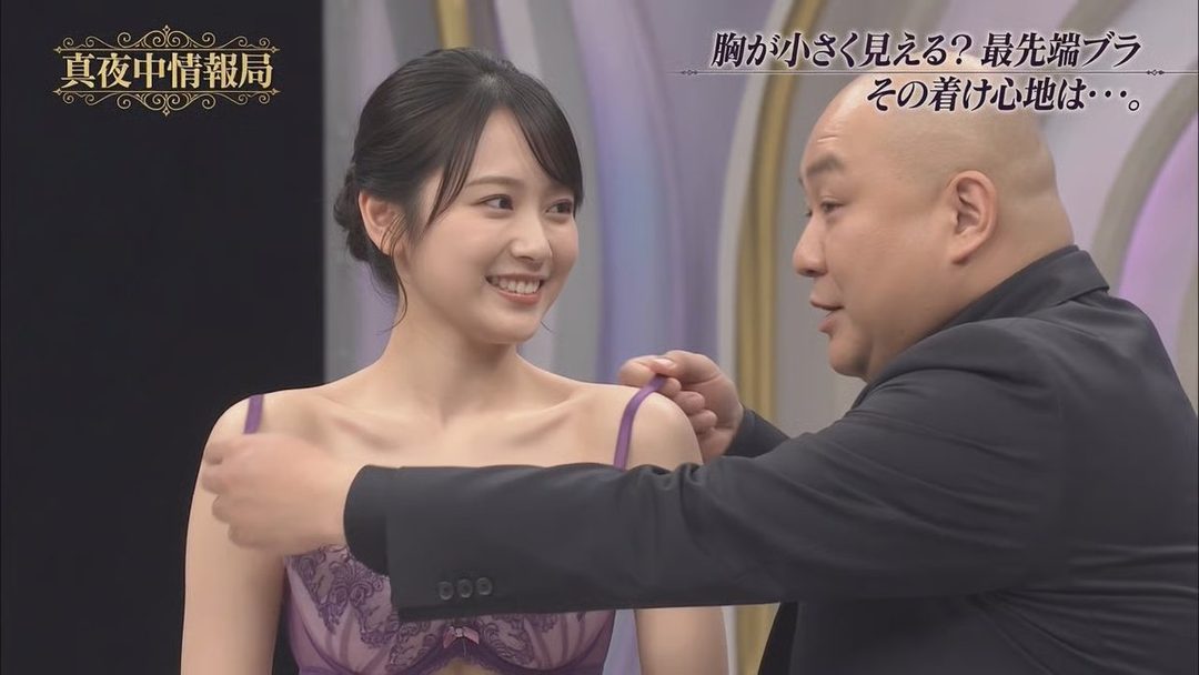 Young Japanese woman in purple lace lingerie smiling while a bald man in a suit adjusts her straps during a late-night TV underwear review segment.