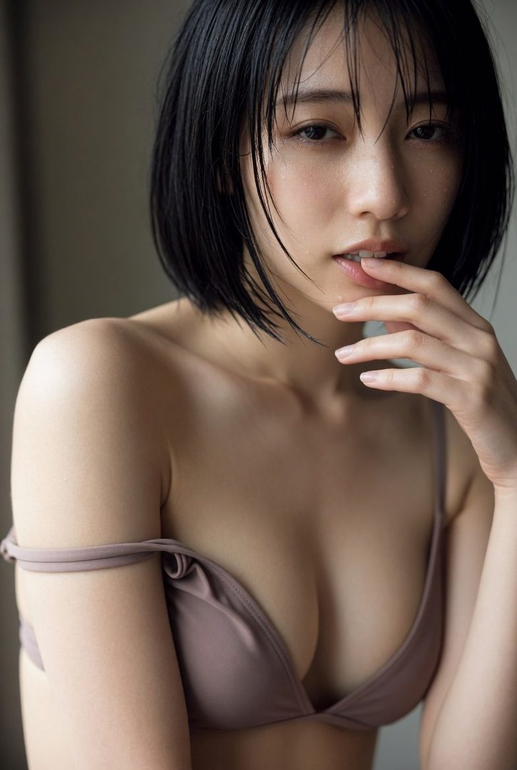 Close-up portrait of a young East Asian woman with short black bob hair wearing a soft mauve bra, posing indoors with hand near her lips.