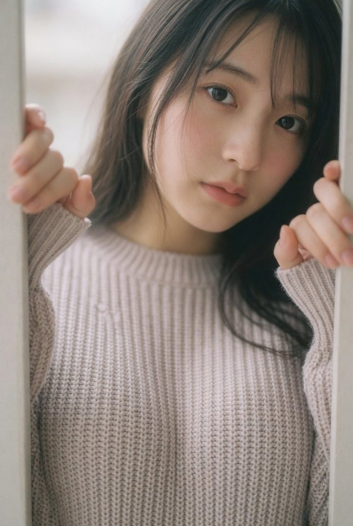 Young East Asian woman with long dark hair and bangs wearing a light pink knit sweater, peeking from behind a door frame while looking at the camera.