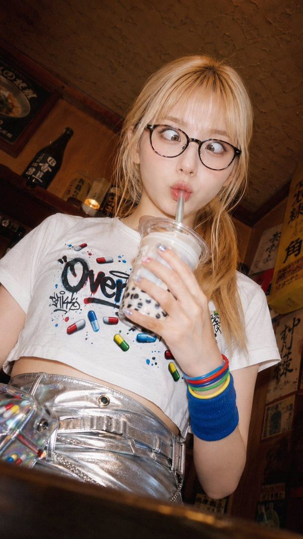 Young blonde woman with glasses sipping bubble tea indoors while wearing a graphic crop top and metallic pants.