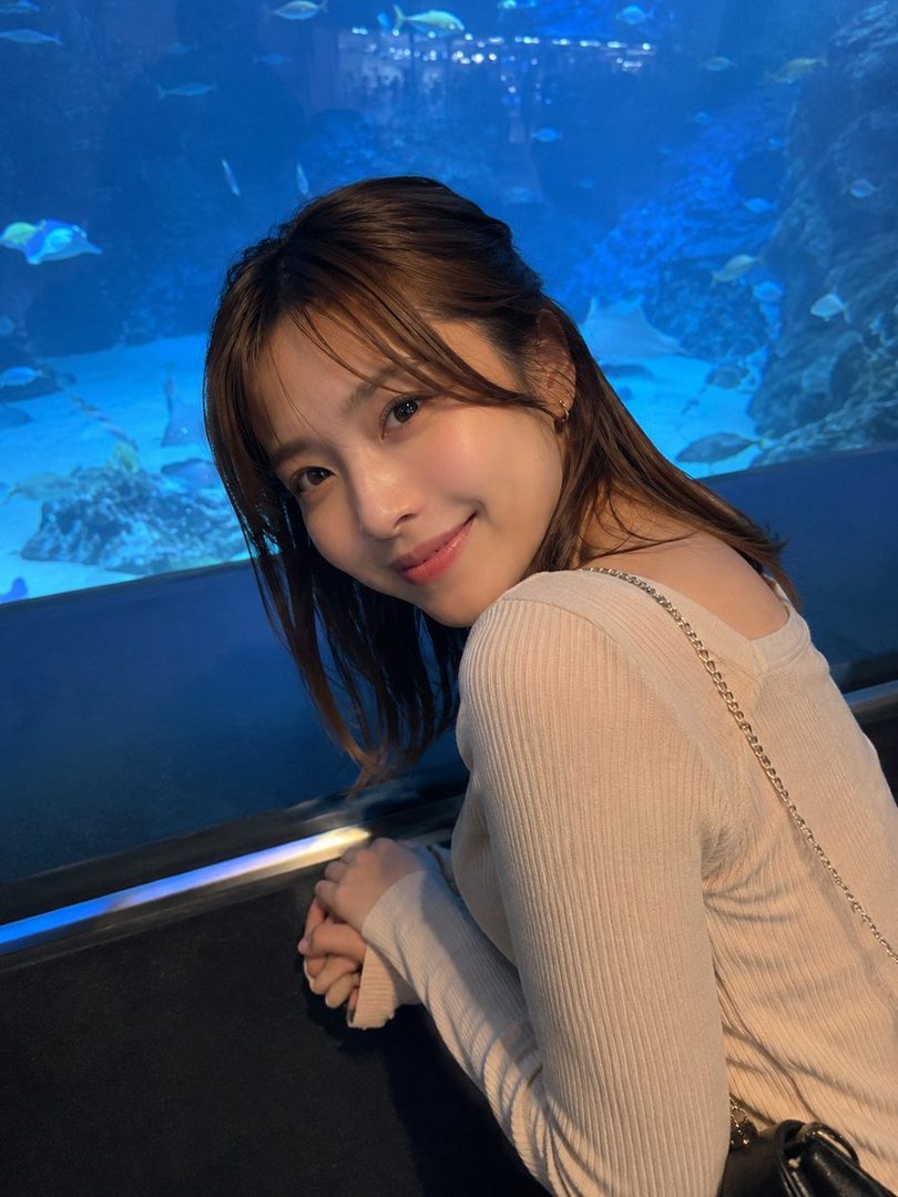 A young woman with brown hair and a ribbed long-sleeve top smiles while looking at fish in an aquarium.