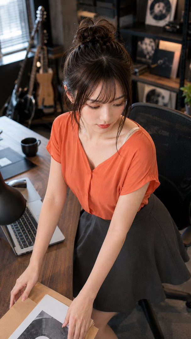 A young Japanese woman with brown hair in a bun wearing an orange blouse and dark skirt examines a vinyl record on a wooden desk in a modern office.