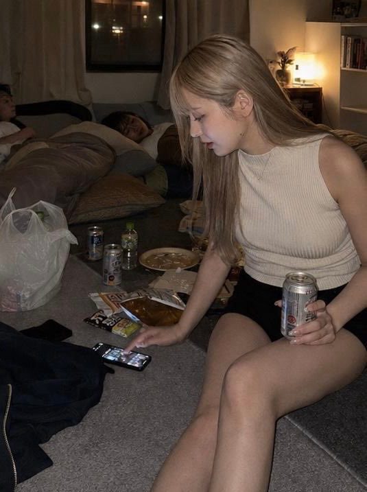 Young blonde woman in casual top and shorts sitting on the floor of a messy living room at night, checking her phone amid snacks and sleeping friends.