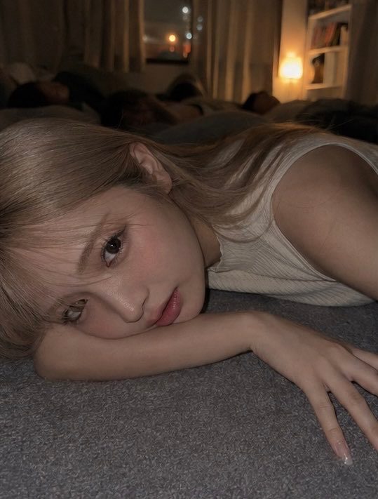 A young blonde woman in a white tank top lies on a carpeted floor in a dimly lit bedroom at night, resting her head on her arm and gazing at the camera.
