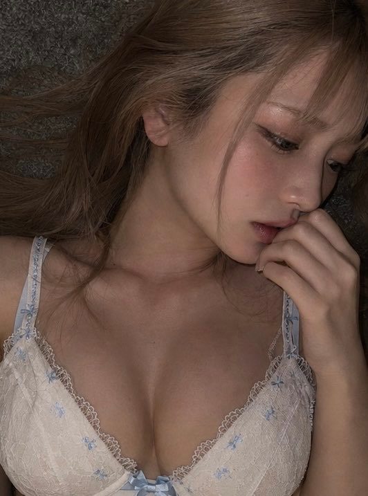 Close-up portrait of a young woman with long light-brown hair lying down in delicate floral lace lingerie, hand near her lips in a soft seductive pose.