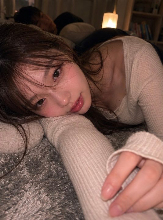 A young woman with long brown hair wearing a beige knit top, lying on a fluffy surface in a cozy indoor room and looking at the camera.