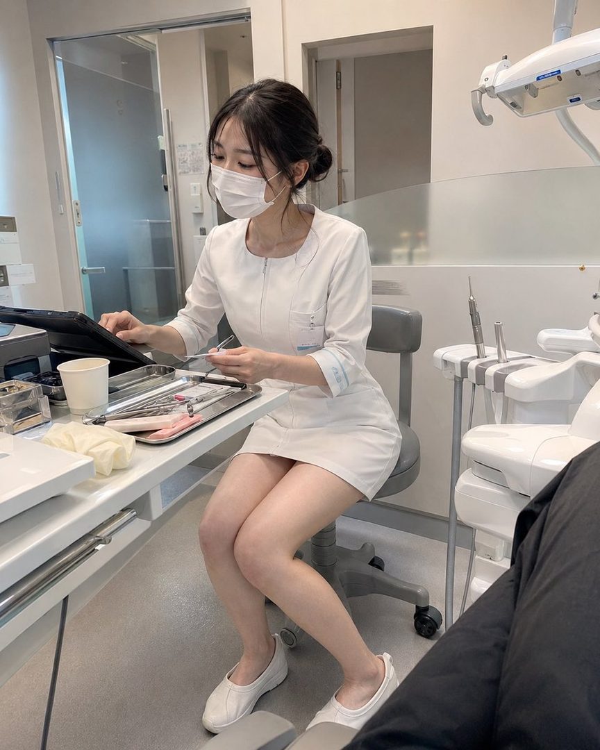 A young East Asian woman in a white medical uniform and face mask sits in a dental clinic holding a clipboard.