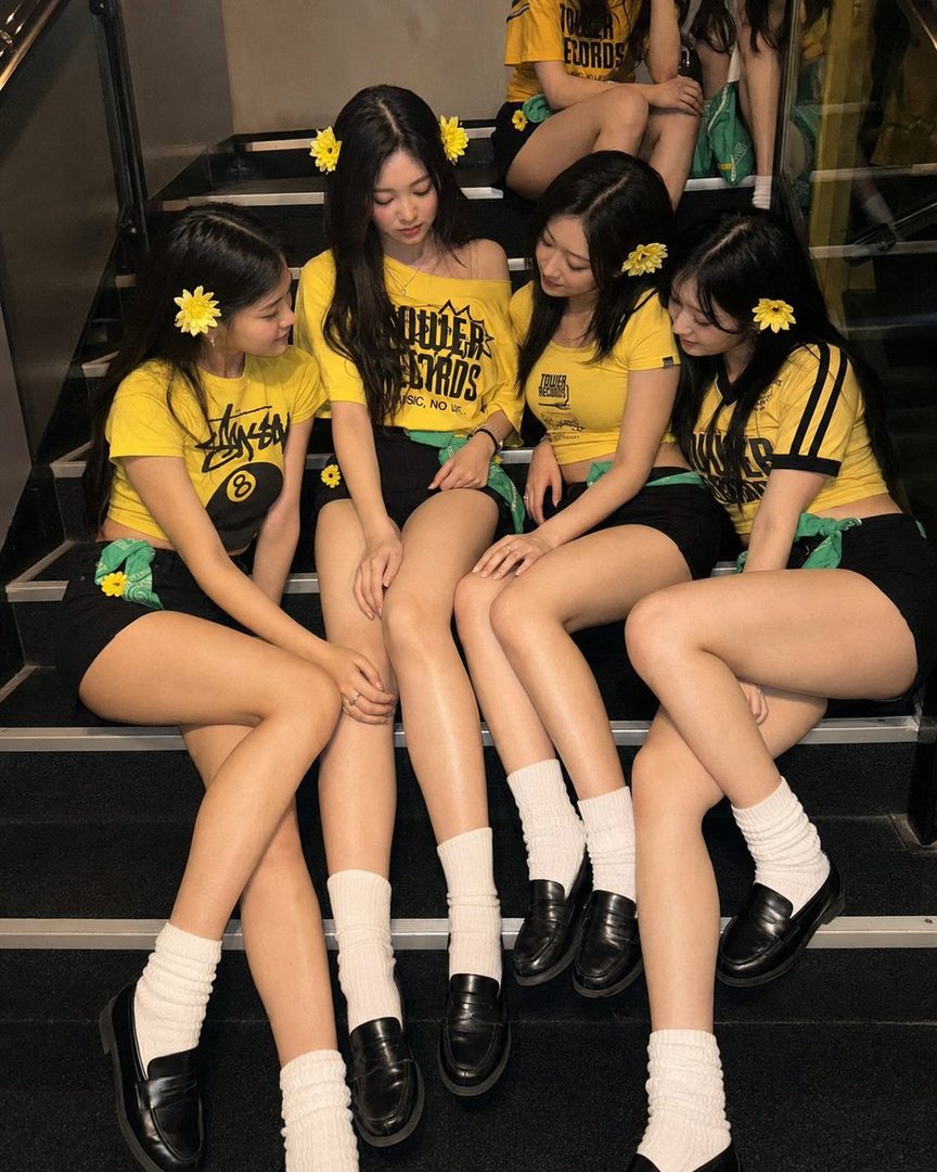 Group of young East Asian women in coordinated yellow graphic tees and black skirts posing together on indoor stairs with yellow flower hair accessories.