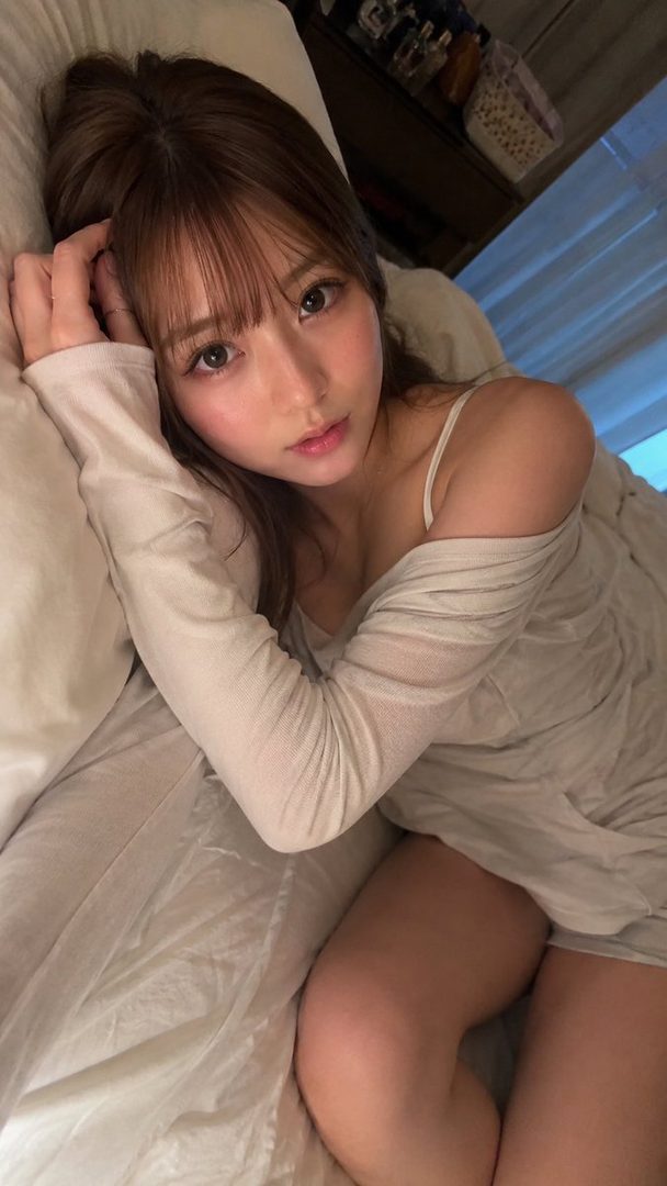 Young East Asian woman with long brown hair and bangs taking a selfie while lying on white bedding in a sunlit bedroom, wearing a sheer off-shoulder top.