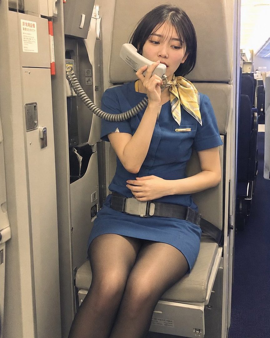Young Japanese flight attendant in blue uniform seated in an airplane cabin holding a phone to her ear.