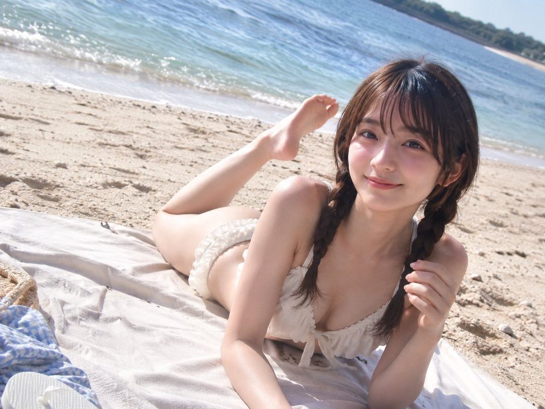 Young East Asian woman in a white bikini posing on a sandy beach with ocean waves behind her.