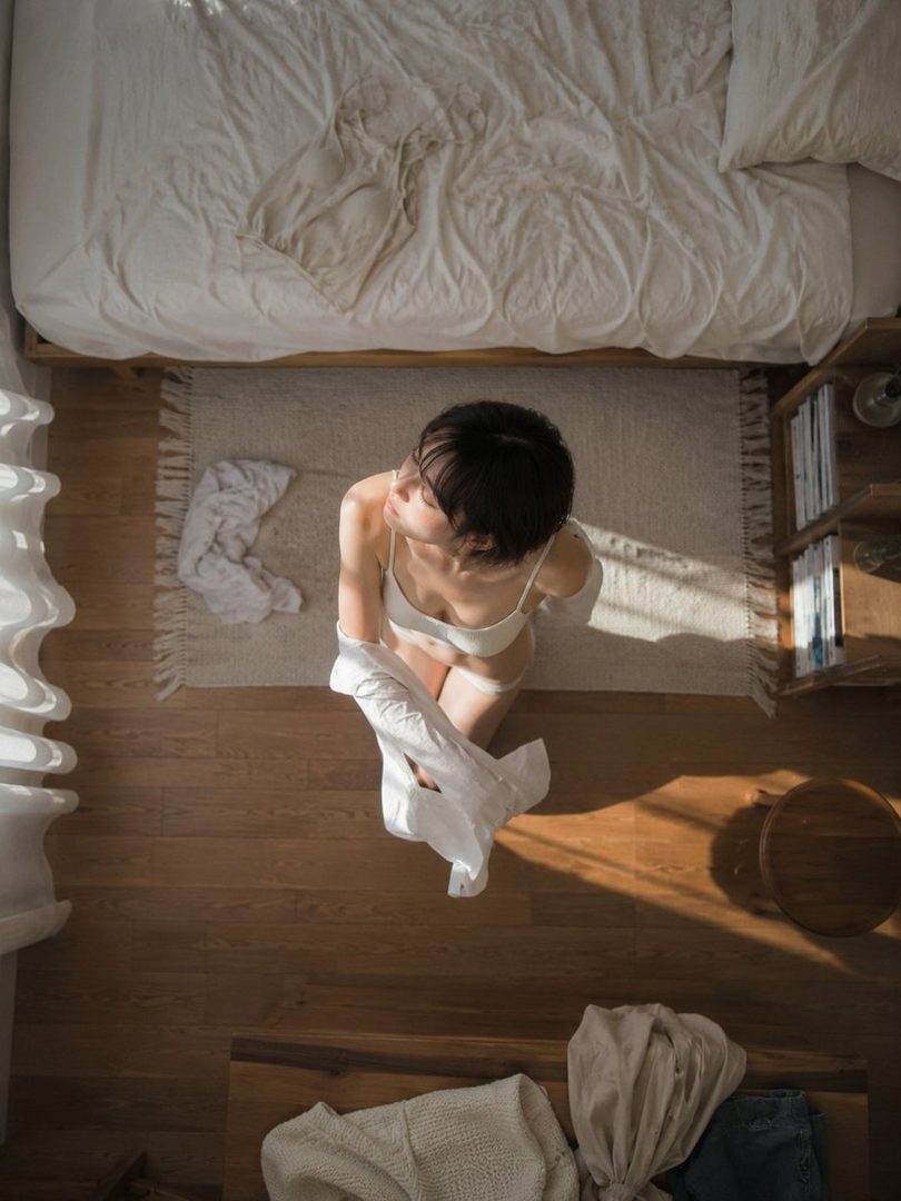 Top-down view of a young woman in white lingerie sitting on a rug in a sunlit bedroom with rumpled white bedding.