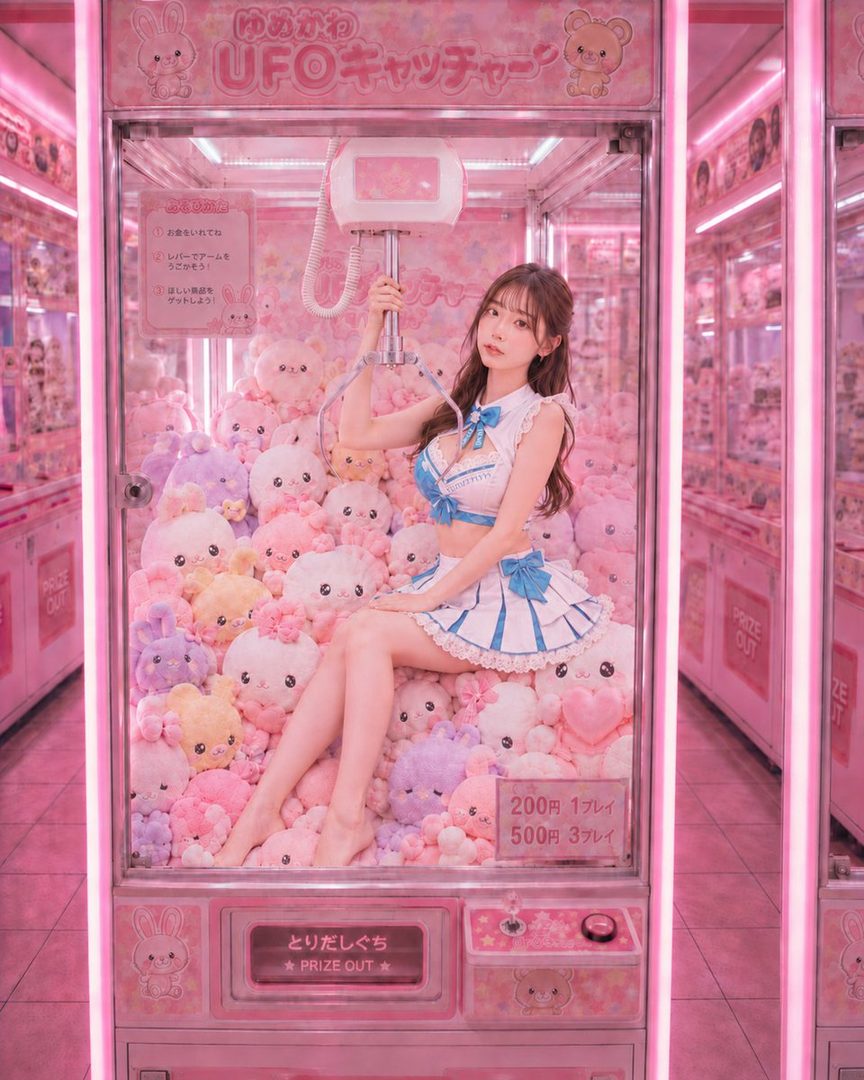 Young woman in a blue and white frilly Alice-style outfit posing inside a pink UFO catcher claw machine filled with plush toys.