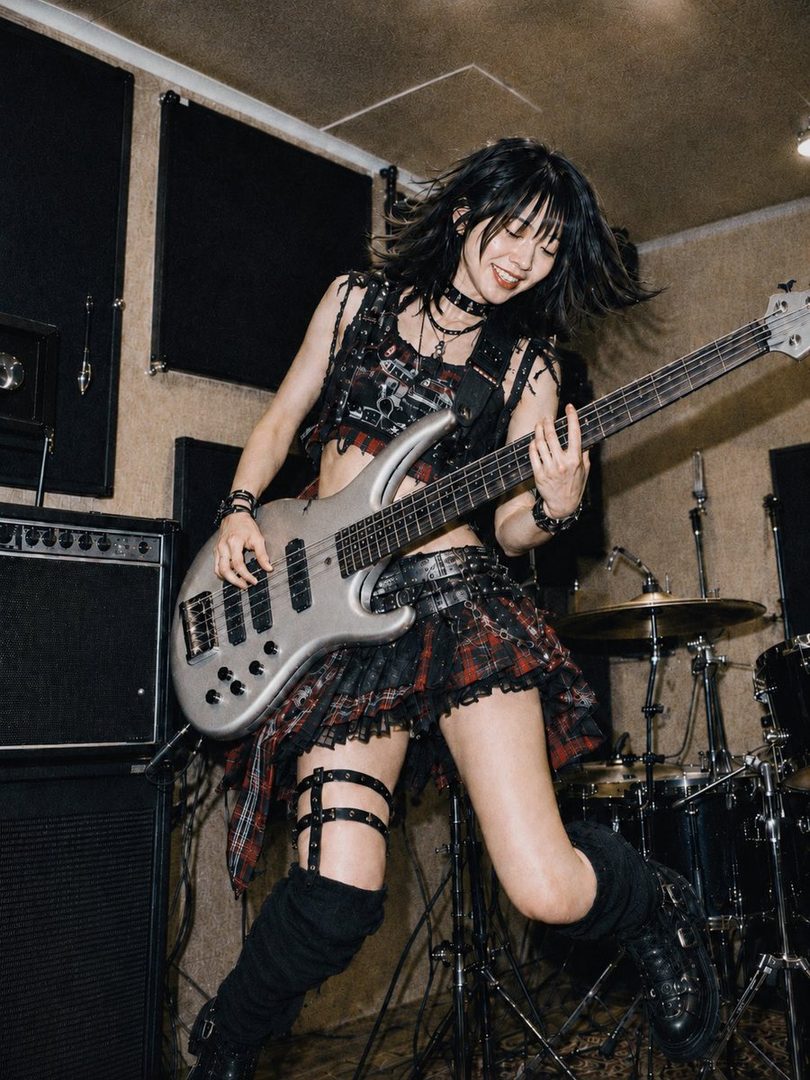 Energetic young woman with messy black bob hair playing a silver bass guitar in a bright Japanese music studio.
