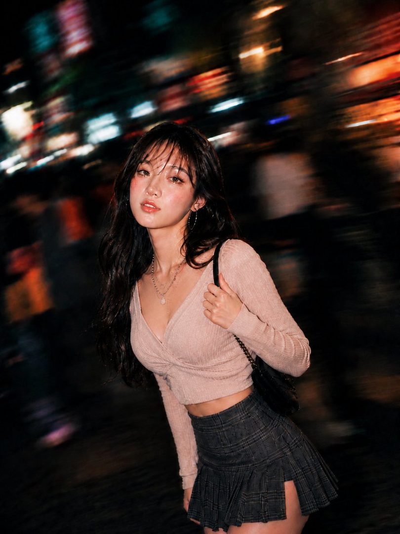 Young East Asian woman with long black hair in a pink crop top and plaid mini skirt posing on a blurred neon night street.