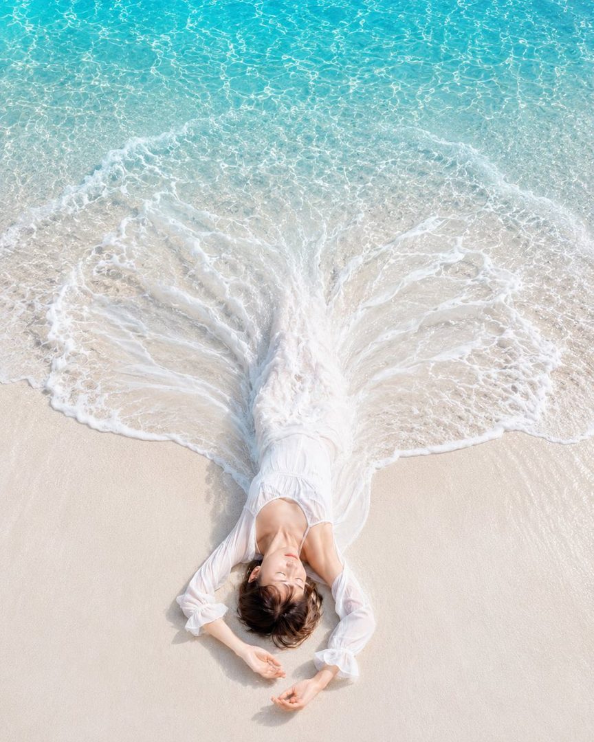 Young Japanese woman with short brown bob hair lying serenely on white sand as turquoise waves flow around her sheer white dress like a flowing hem.