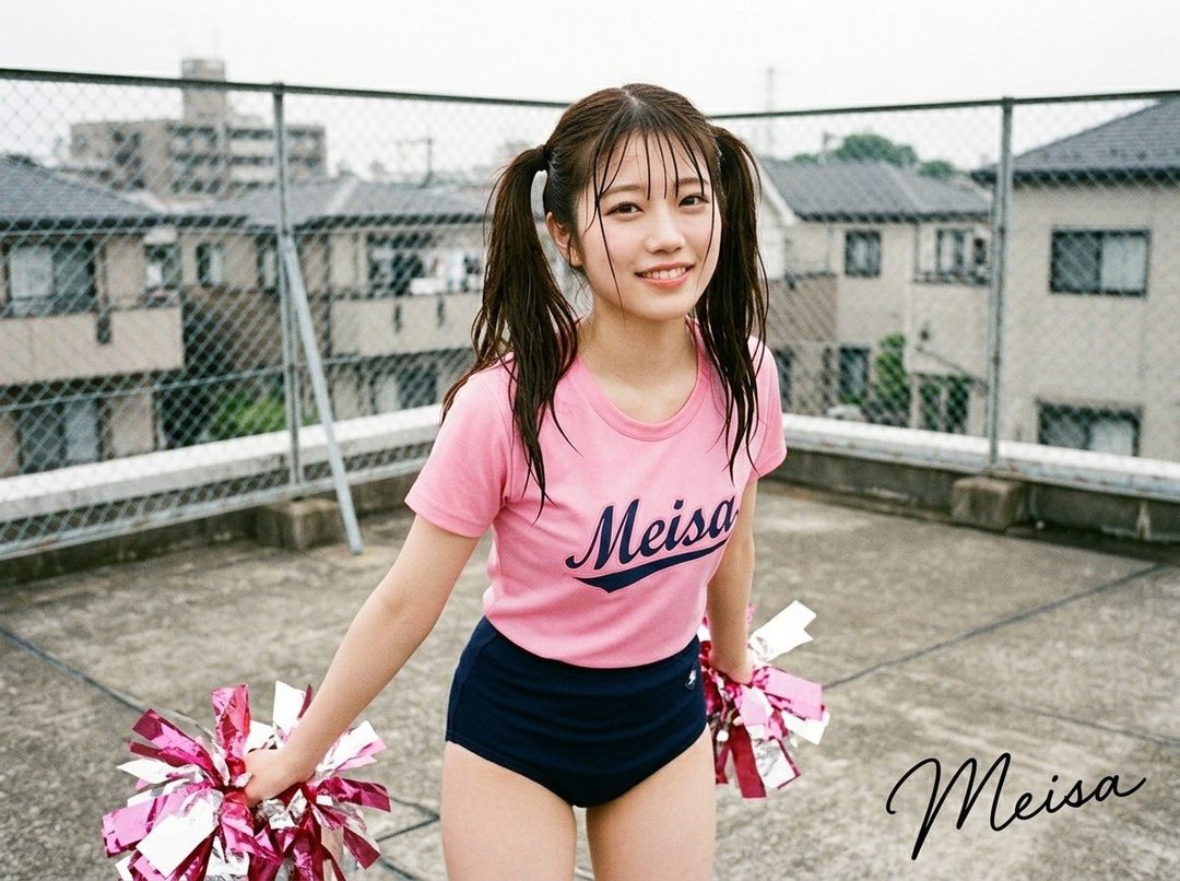 Smiling young woman with brown twintails in a pink t-shirt and navy bloomers holding pink pom-poms on an urban rooftop.