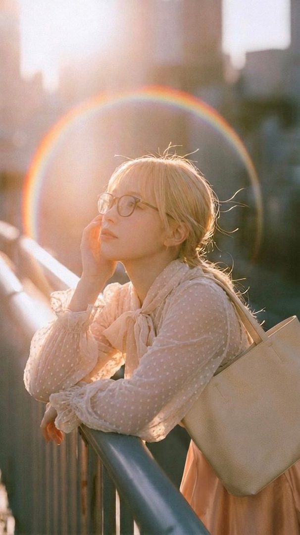 A blonde woman with glasses leans on a railing, looking thoughtfully into the distance with a soft lens flare and rainbow effect in the background.
