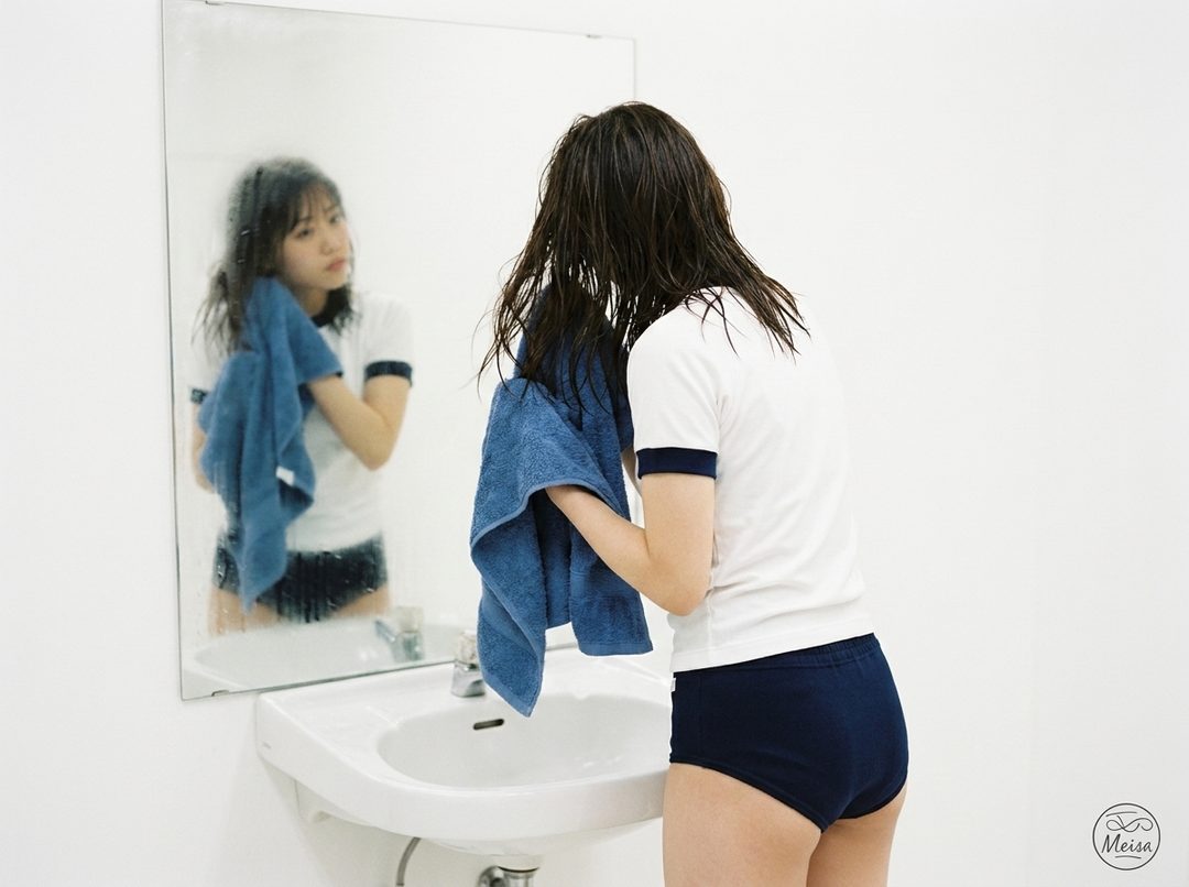 Young woman with wet dark hair in gym uniform drying off in a bright white bathroom, viewed from behind with mirror reflection.