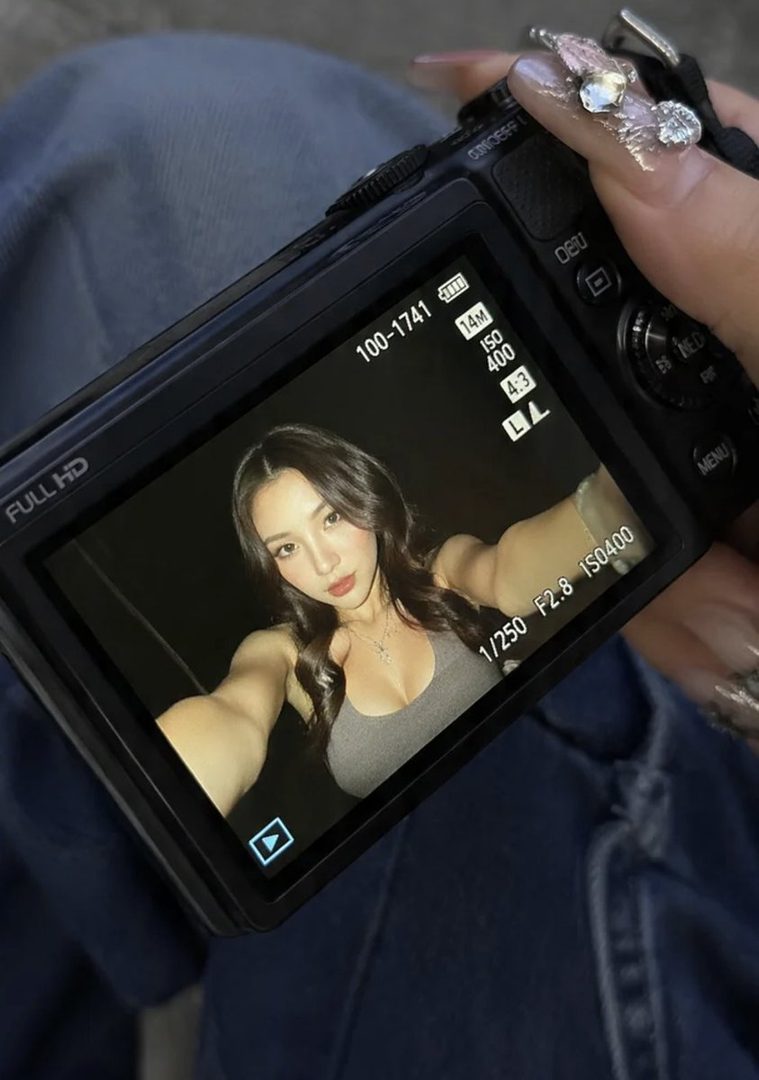 Young woman taking a mirror selfie on a digital camera screen, wearing a gray tank top with long dark hair.