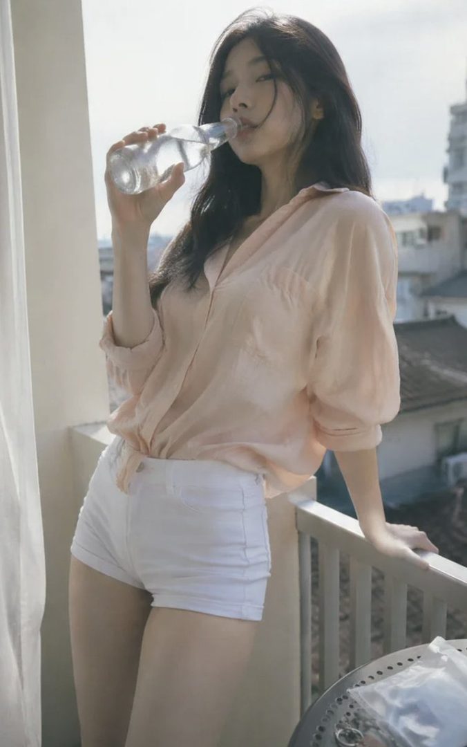 A young East Asian woman with long black hair in a light pink blouse and white shorts drinks from a bottle on a sunny city balcony.