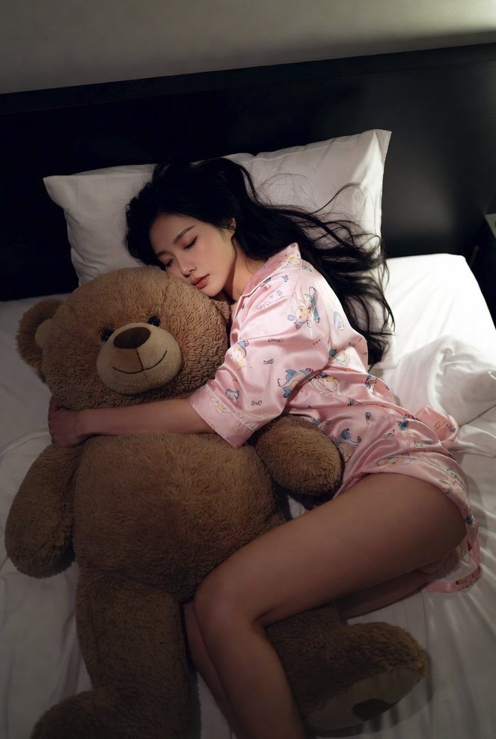 Young woman with long black hair in pink pajamas hugging a large teddy bear while lying in bed.