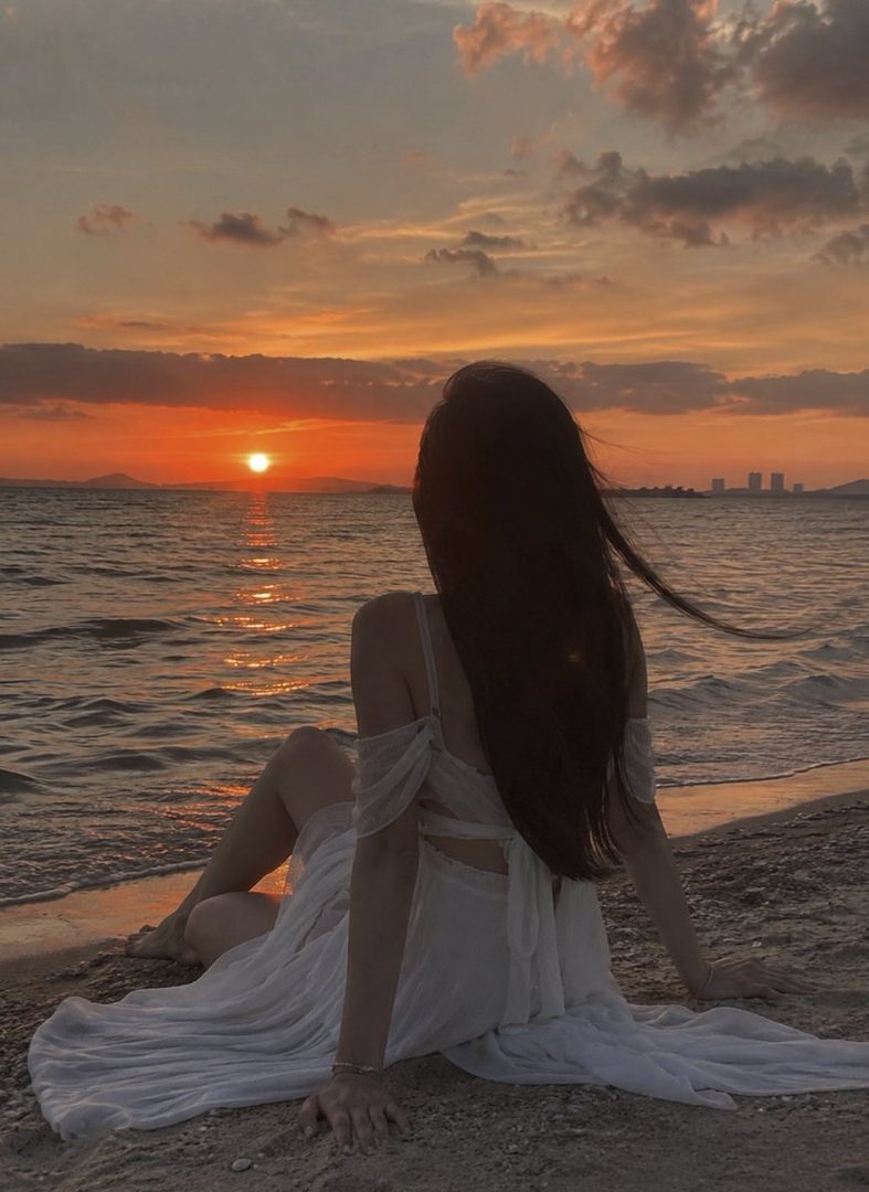 A woman with long dark hair wearing a flowing white dress sits on the sand gazing at a vibrant ocean sunset.