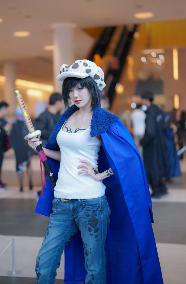 Woman cosplaying as Trafalgar Law from One Piece at an indoor anime convention, wearing a spotted hat, blue coat, and holding a sword.