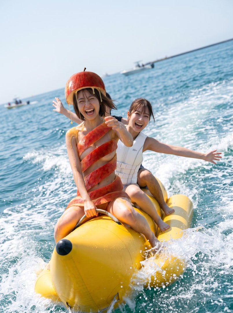 Two young women laughing on a yellow banana boat in the ocean, one wearing a playful watermelon costume.