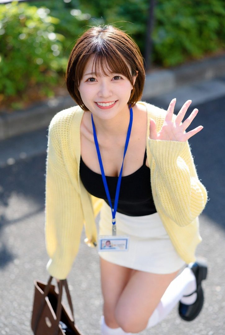 Smiling young woman with short brown bob hair in a yellow cardigan waves cheerfully outdoors on a sunny day.